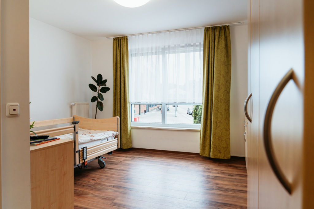Bewohnerzimmer mit Pflegebett, großem Fenster und Vorhängen