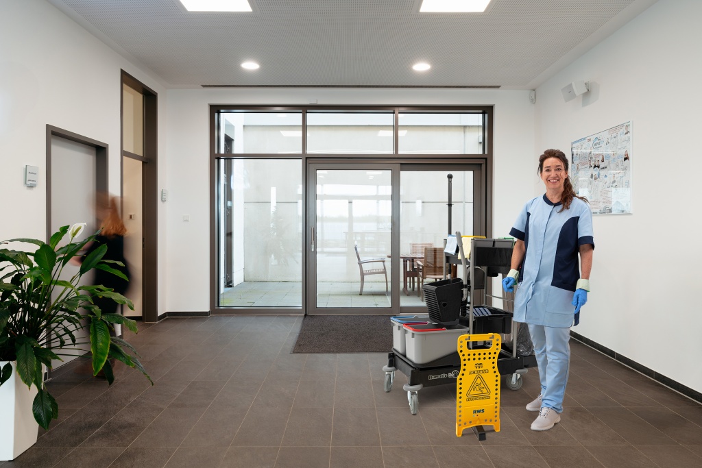Mitarbeiterin von RWS Gebäudeservice steht mit Reinigungswagen und Hinweisschild im Eingangsbereich einer Verwaltung