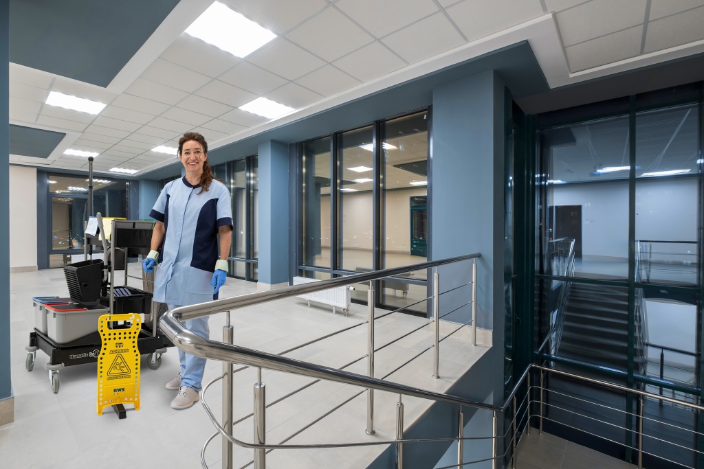 Mitarbeiterin von RWS Gebäudeservice steht mit Reinigungswagen und Warnschild im Treppenhaus eines modernen Bürogebäudes.