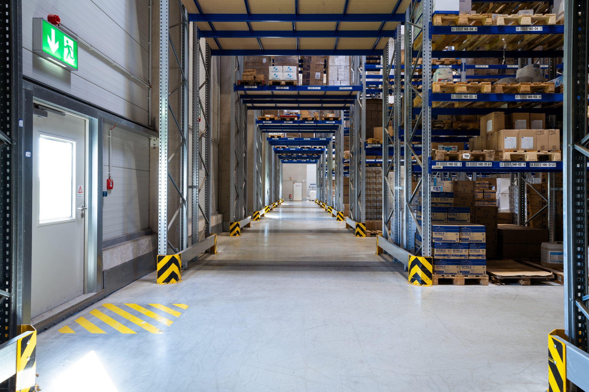 Sauberer Lagergang mit hohen Regalen, klar markierten Bodenflächen und einer Notausgangstür.