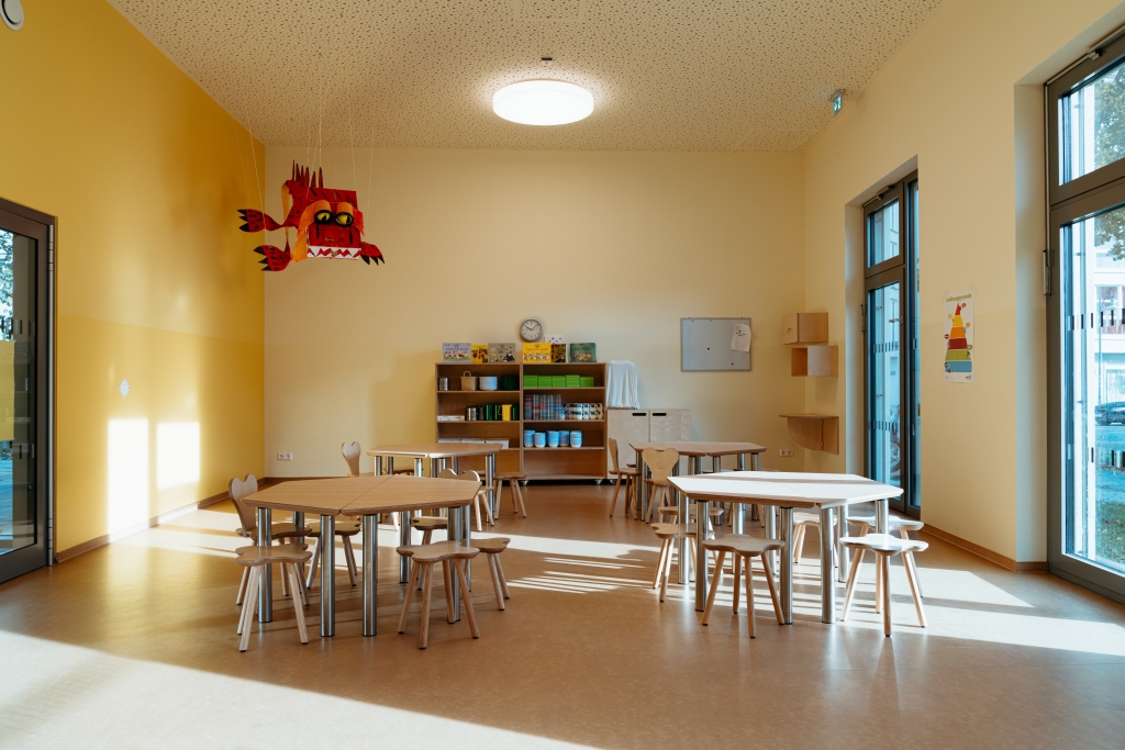 Heller Gruppenraum in einer Kita mit Tischen, Stühlen und bunter Dekoration