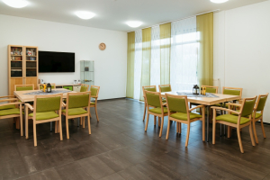 Gemeinschaftsraum mit Tischen, Stühlen und Fernseher im Pflegeheim