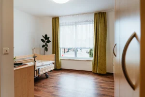 Bewohnerzimmer mit Pflegebett, großem Fenster und Vorhängen