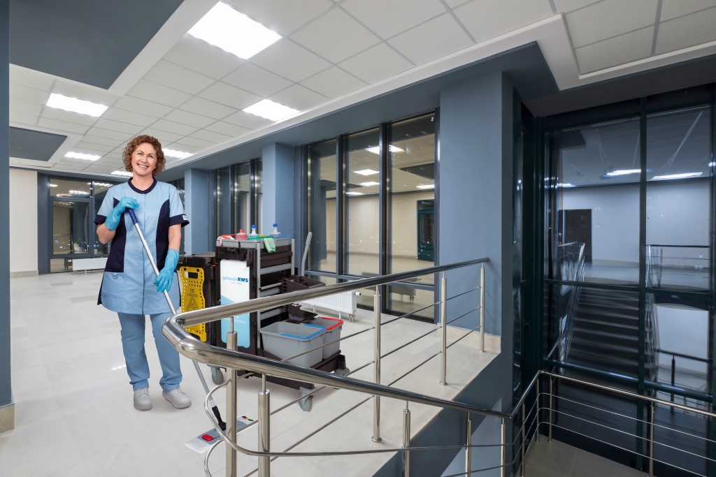 Mitarbeiterin von RWS Gebäudeservice steht mit Reinigungswagen und Warnschild im Treppenhaus eines modernen Bürogebäudes.